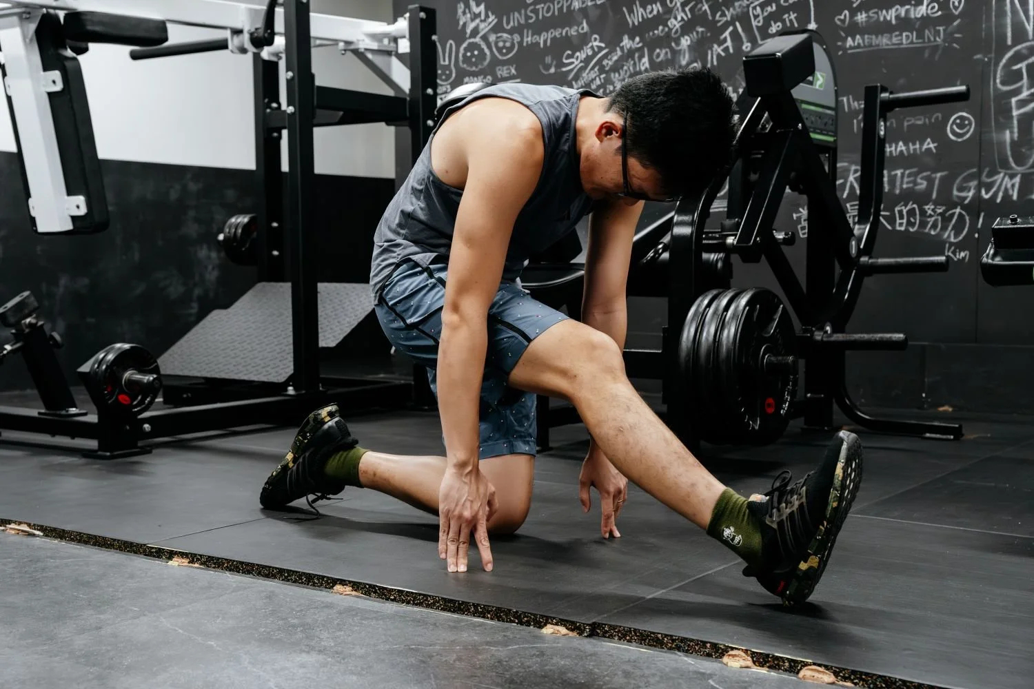 A man wearing a sleeveless shirt, shorts, and glasses is stretching on the gym floor, with gym equipment and a black chalkboard wall with writings behind him.
