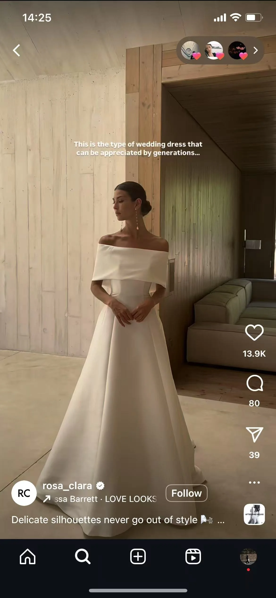 A woman in an off-the-shoulder white wedding dress standing indoors against light wood-paneled walls and a gray couch.