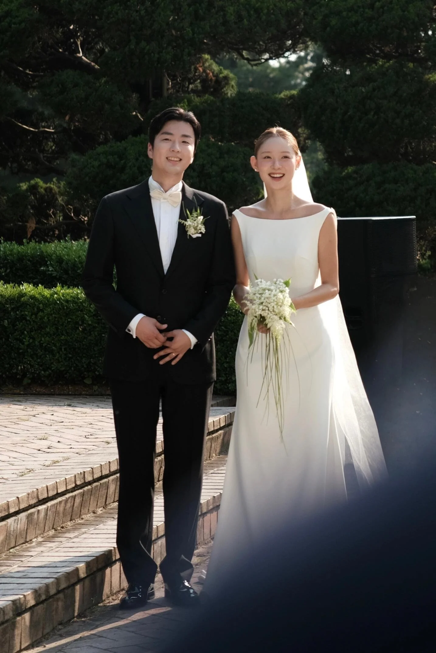 A bride and groom standing outdoors on a walkway, smiling. The groom wears a black tuxedo with a beige bow tie and a small floral boutonniere. The bride wears a white wedding gown, holds a bouquet of white flowers, and has her hair styled back with a veil. Behind them is greenery and trees, with sunlight filtering through.
