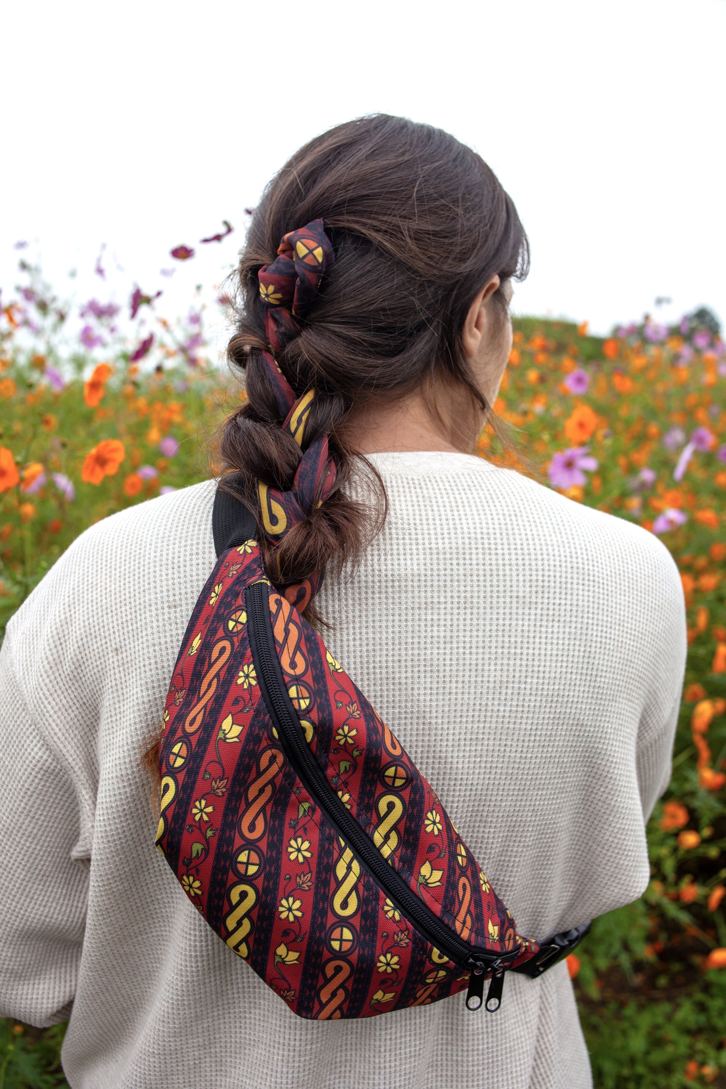 Back view of a woman with braided hair, wearing a cream sweater, standing outdoors in a field of orange, pink, and purple flowers, with a colorful patterned bandolier bag slung over her shoulder.