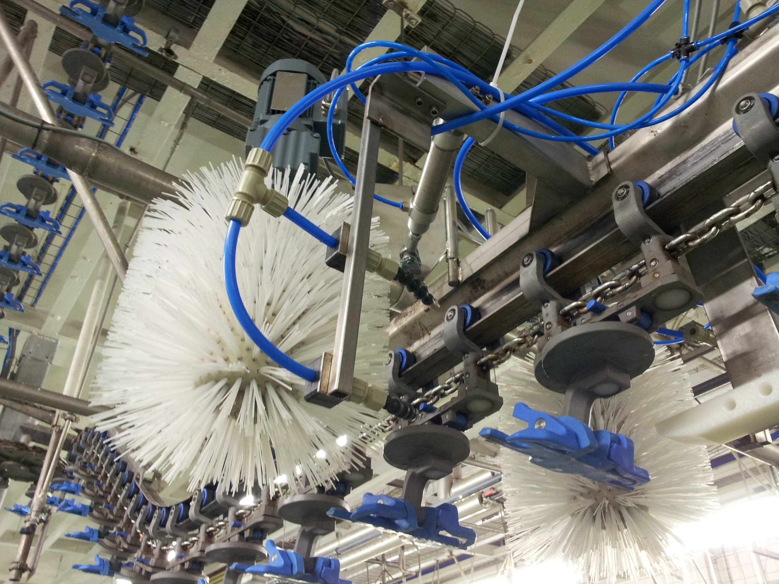 Industrielle automatische Reinigungsmaschine mit weißen Bürsten, blauen Rohren und Metallrahmen in einer Fabrikhalle.