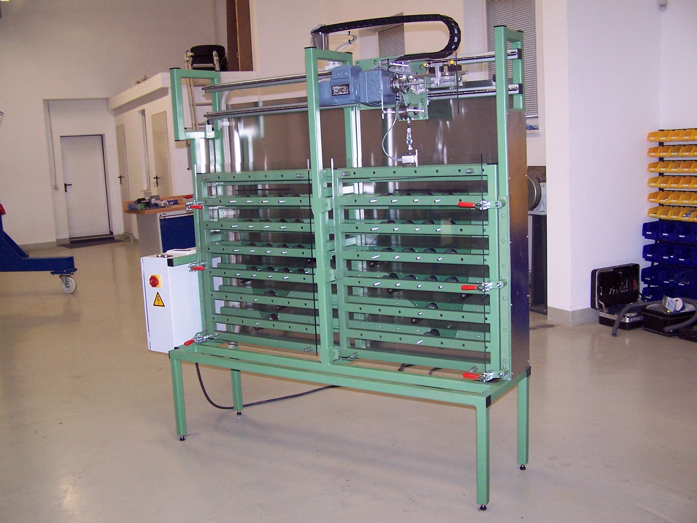 Industriegerät mit grüner Metallrahmenkonstruktion, mehreren horizontalen Schienen und einer Steuerbox mit rotem Not-Aus-Button in einem Werkstattraum.