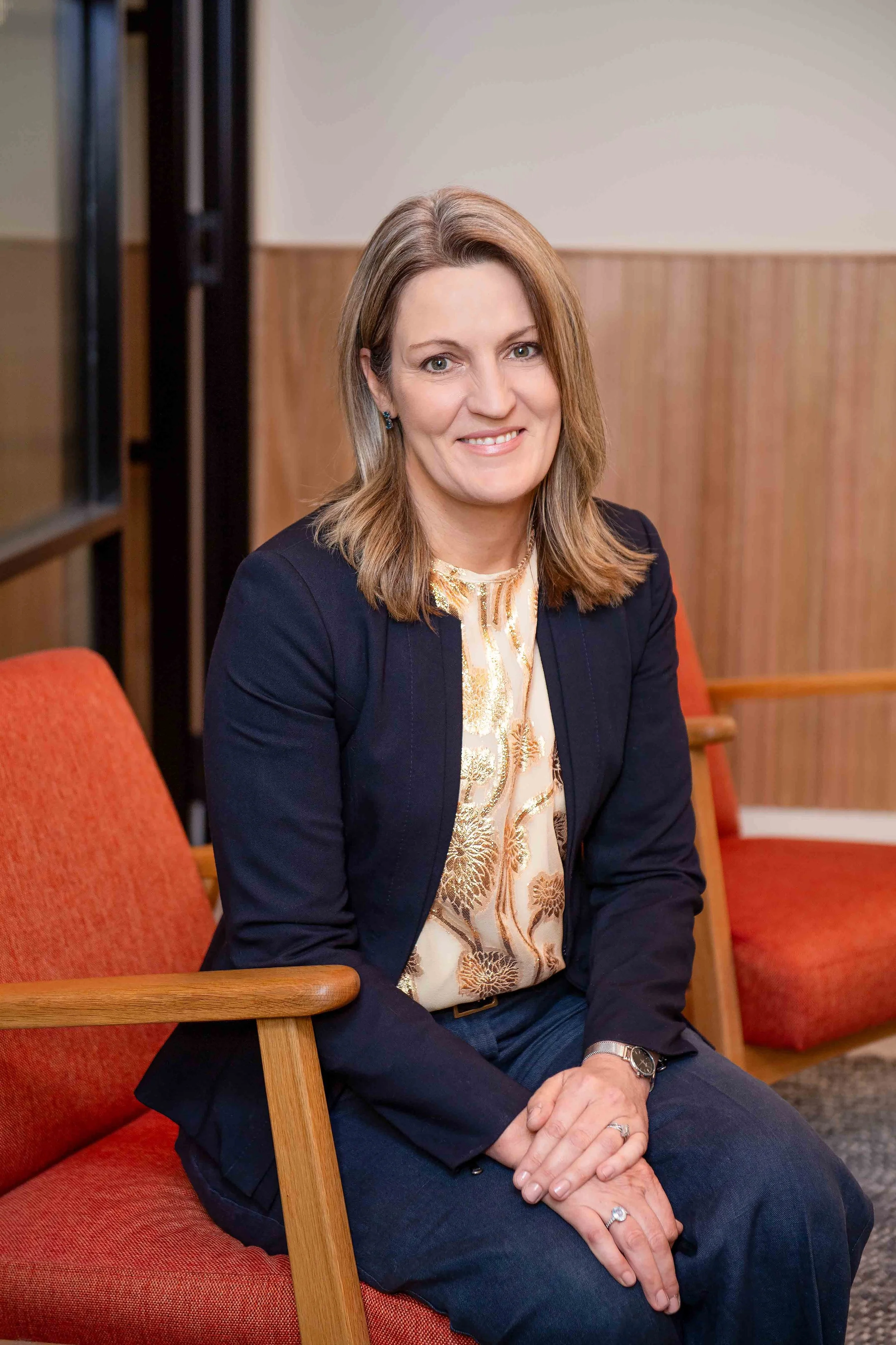 A woman sitting on an orange upholstered chair in a room with wood-paneled walls, wearing a black blazer, light patterned blouse, dark jeans, and a watch, smiling at the camera.