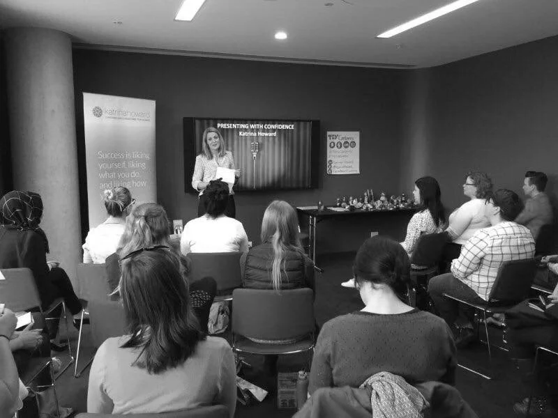 A woman giving a presentation to a seated audience in a conference room, with a large screen behind her displaying the title 'Presenting with Confidence' and her name, Karina Howe.