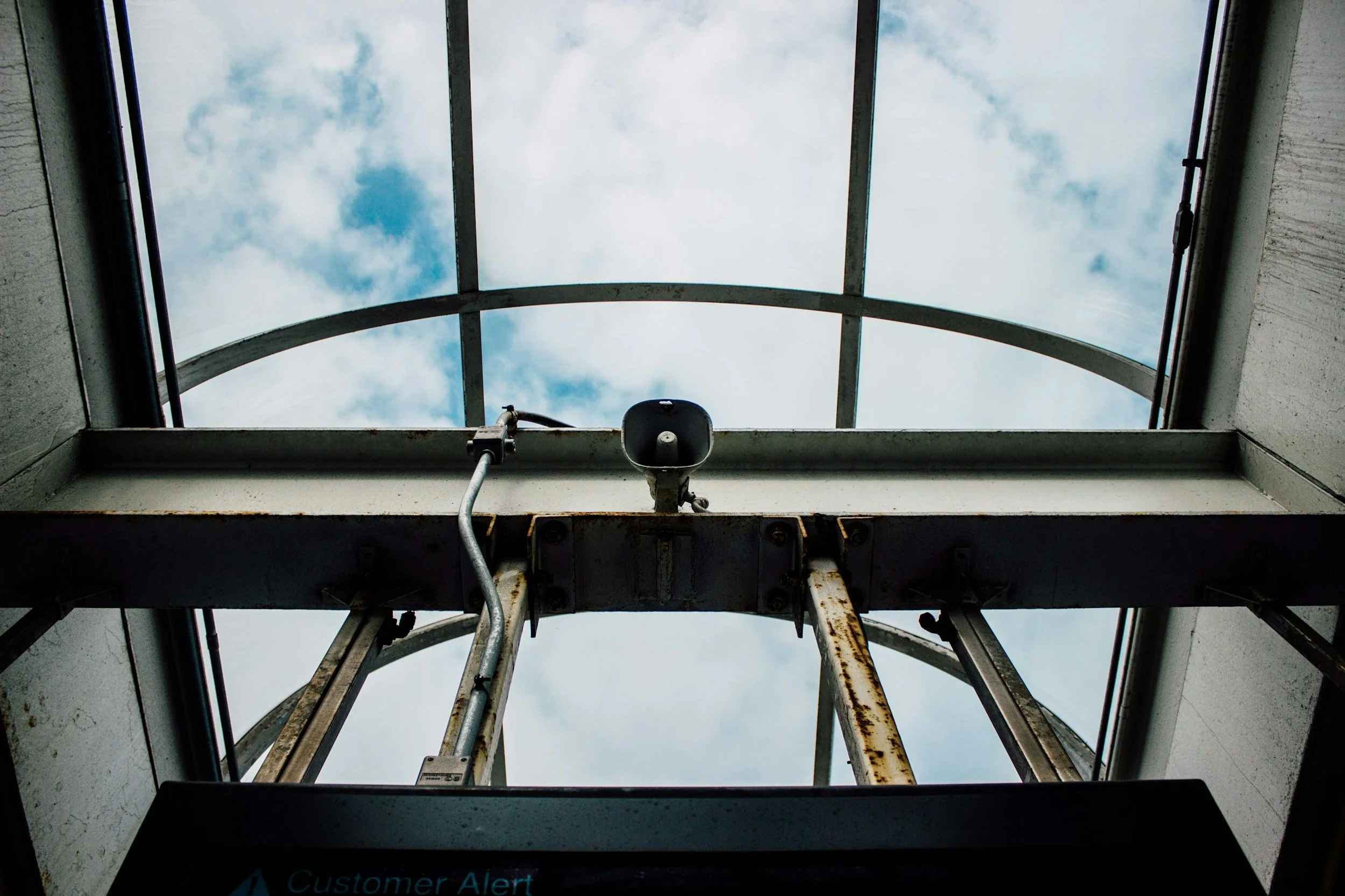 View looking up at an open skylight with a cloudy sky above. The skylight has a metal frame and an exposed speaker or alarm attached to the structure below the glass.