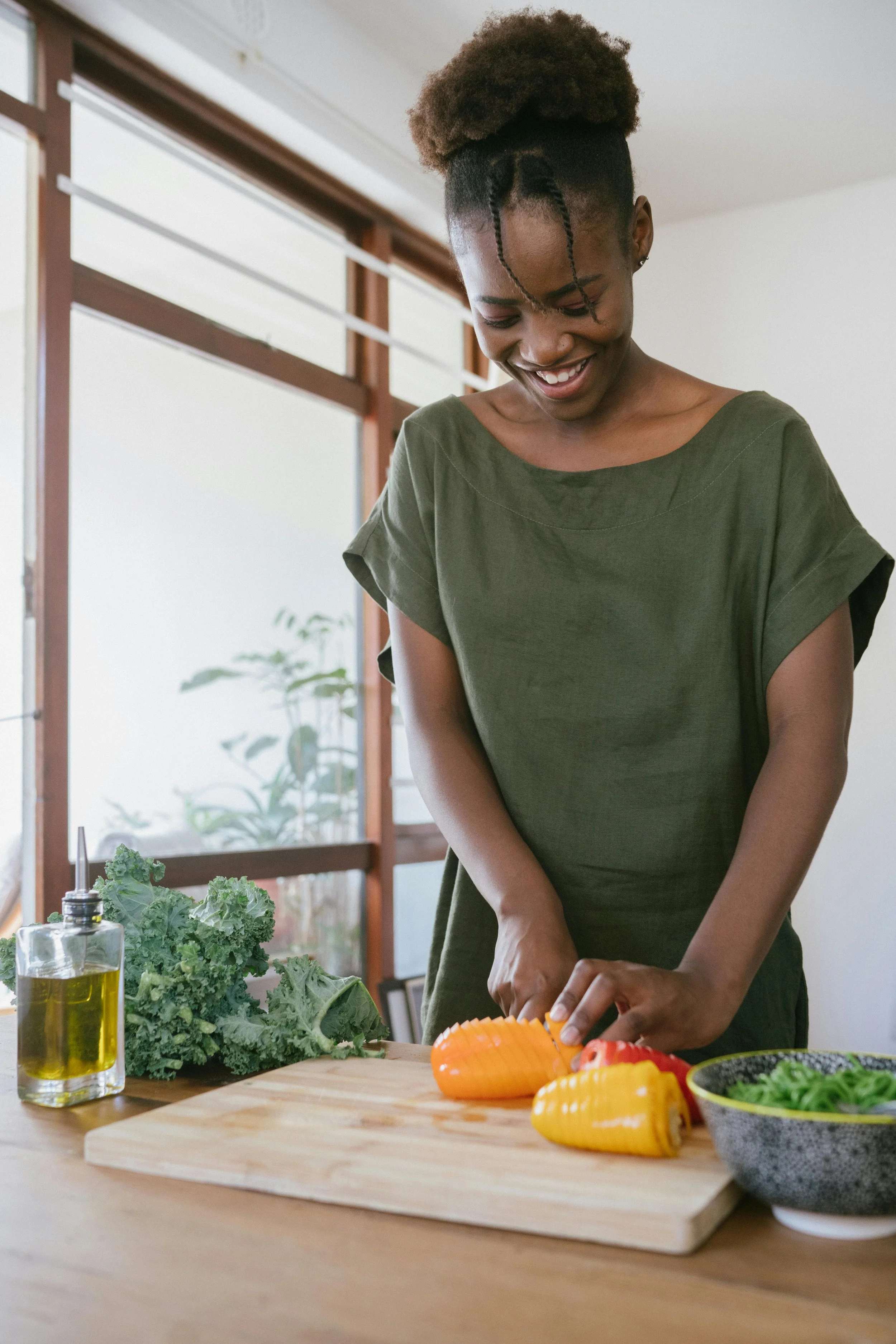 A woman smiling while slicing a yellow bell pepper in a bright kitchen with large windows, next to a bowl of chopped green vegetables, a head of kale, and a bottle of olive oil on the wooden table.
