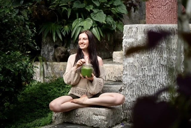 A woman sitting cross-legged on a stone step outdoors, holding a green drink, smiling amidst lush greenery.