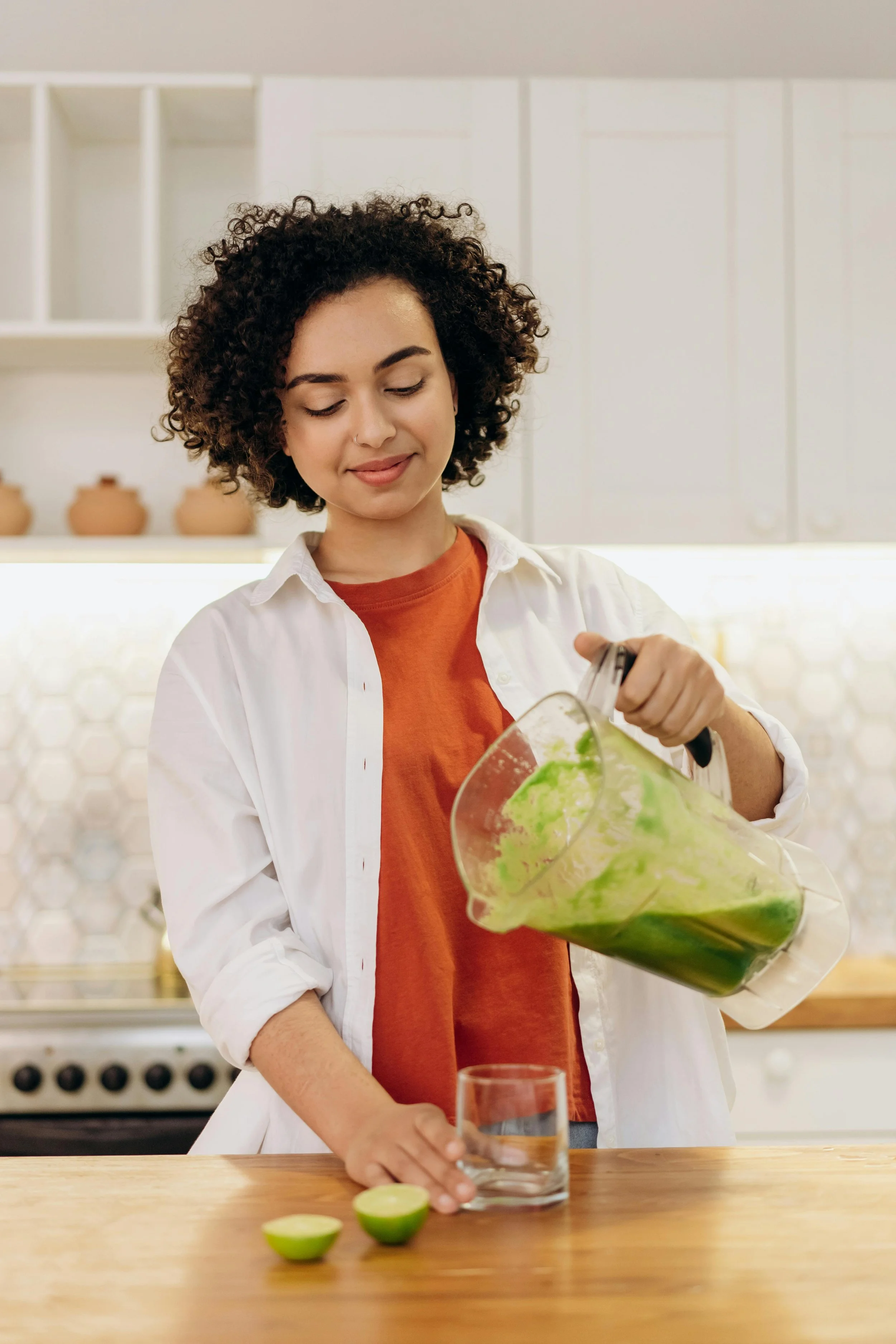 A woman in a white shirt and orange t-shirt is pouring a green-colored smoothie from a blender into a glass on a wooden countertop.