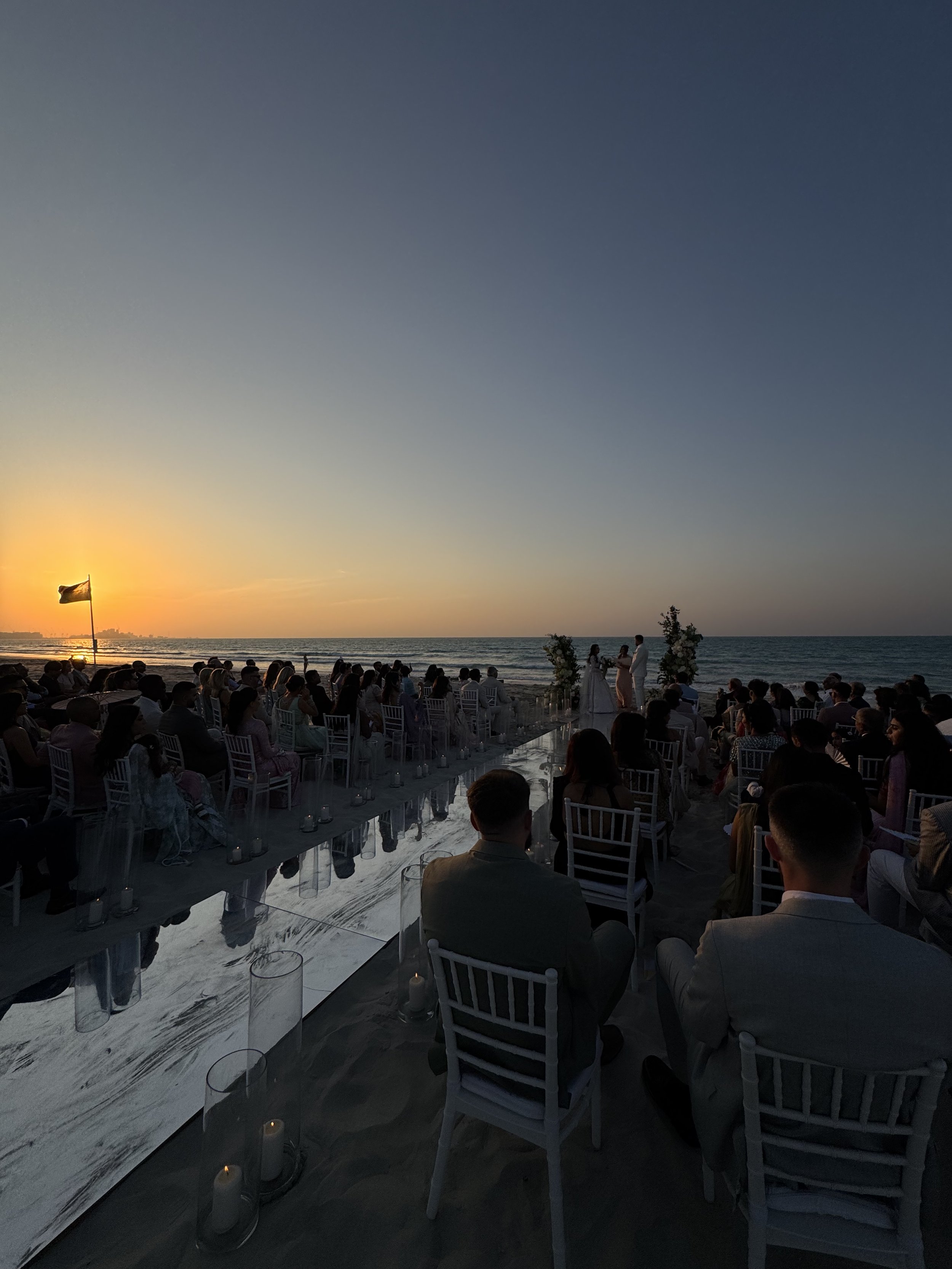 Wedding ceremony on a beach at sunset with guests seated and an officiant at the altar, ocean in the background.