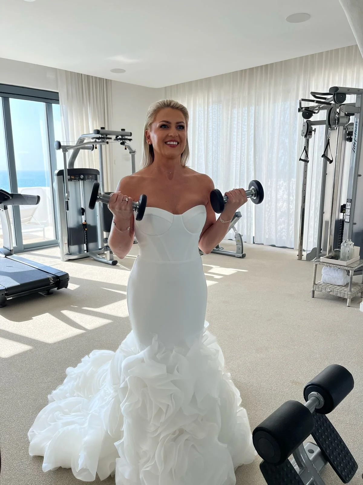 A woman wearing a white wedding dress lifts dumbbells in a gym with large windows showing an ocean view.