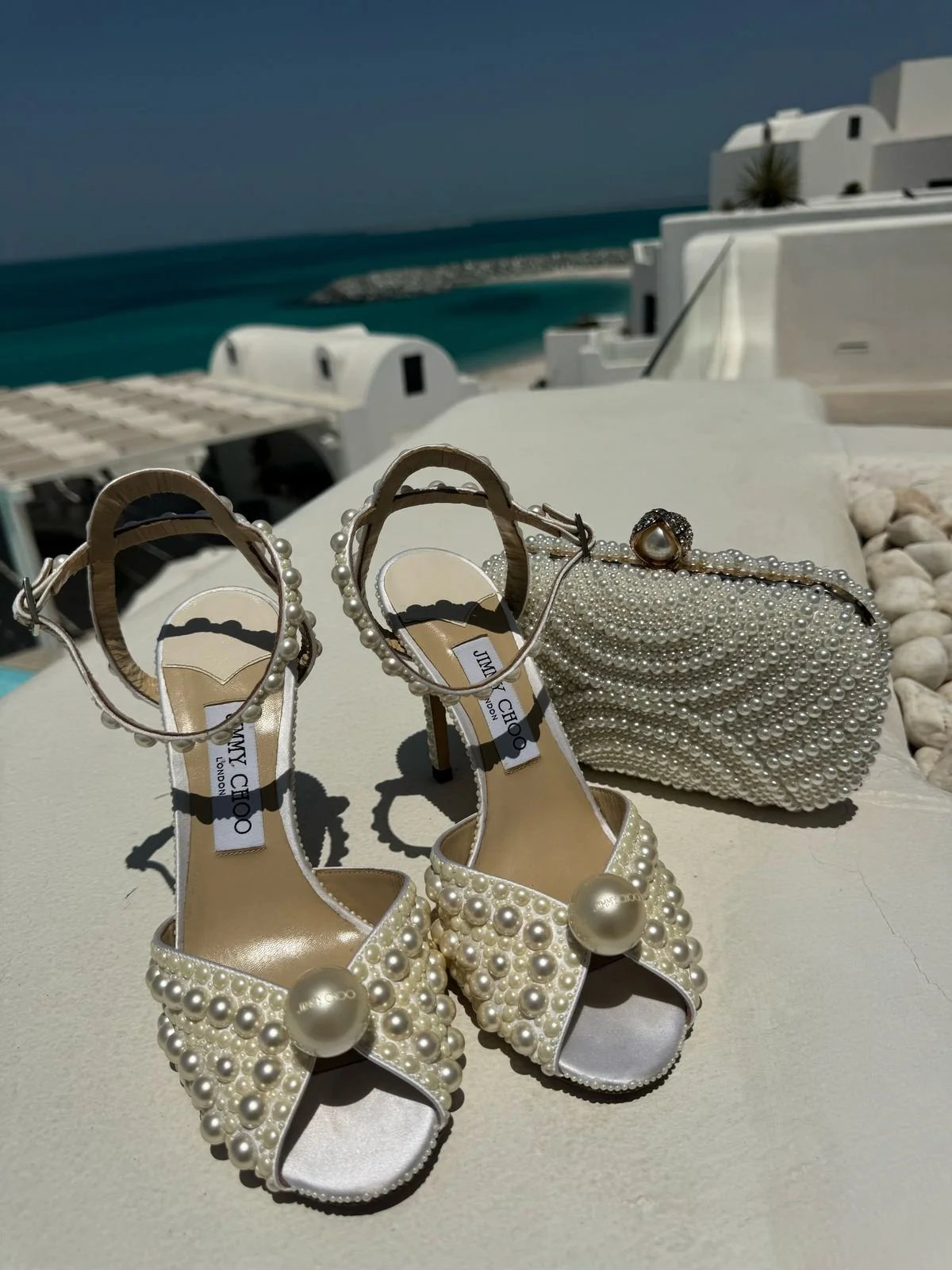Pearl-embellished high-heel shoes and a pearl clutch purse placed on a white surface under natural sunlight with a scenic coastal background featuring white buildings and blue sea.