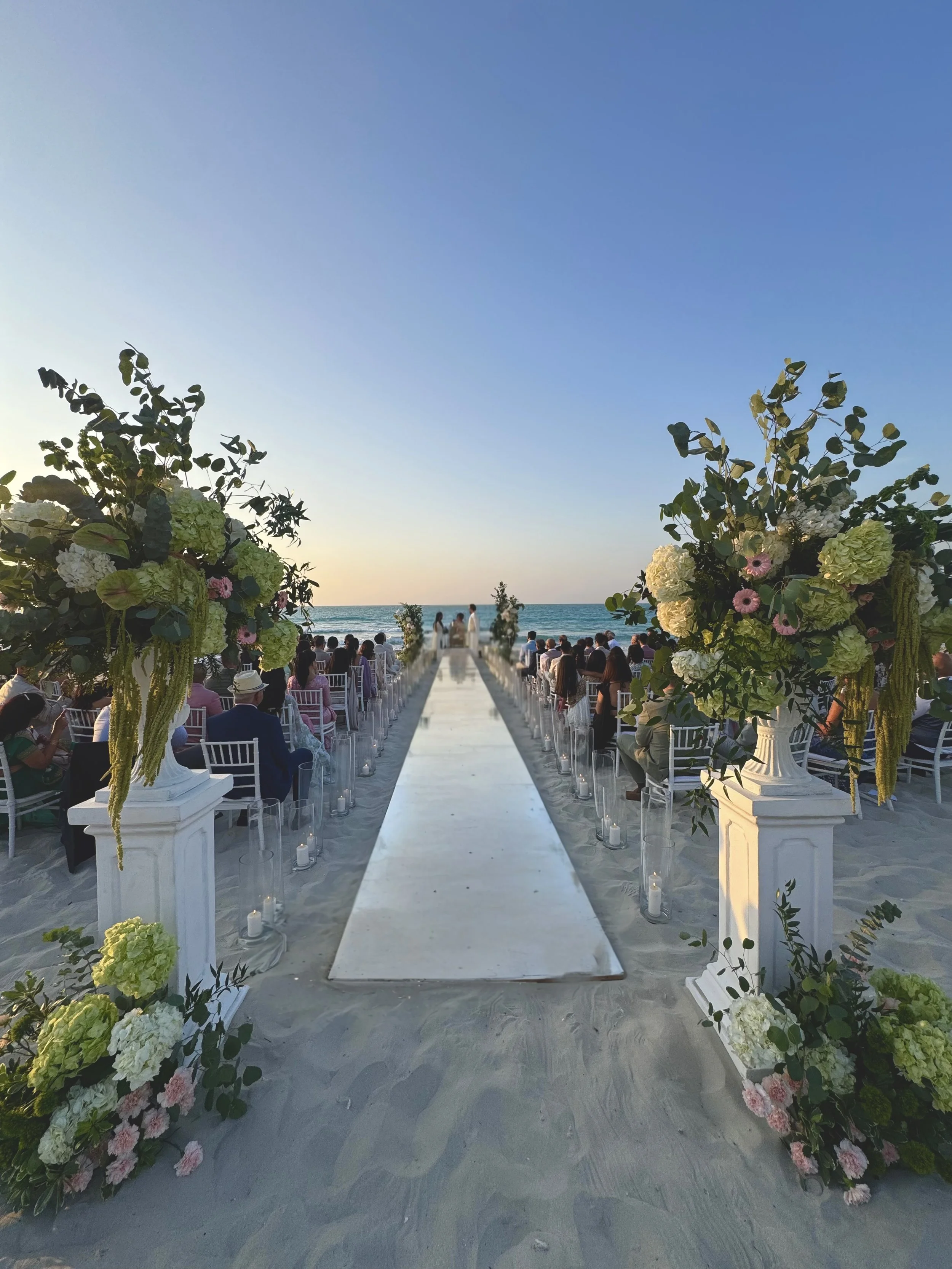 Beachside wedding ceremony with floral arrangements, candles, and chairs facing the ocean at sunset.