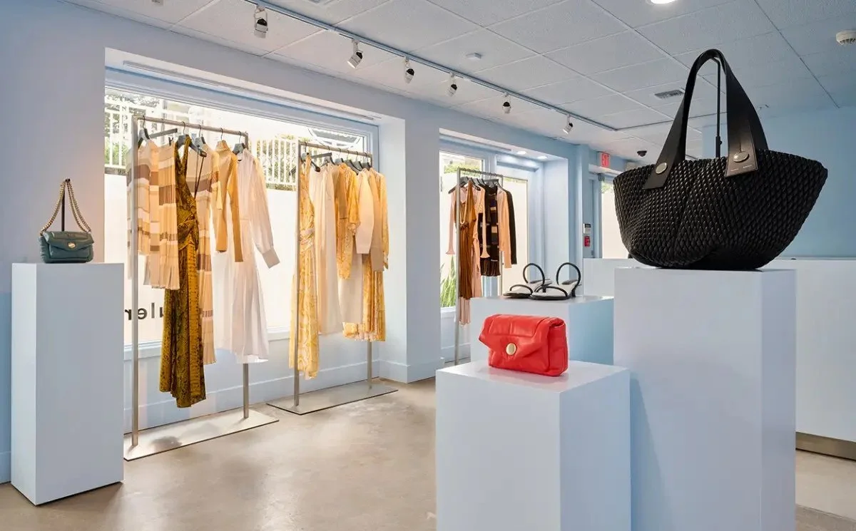 Clothing store with pastel blue walls, featuring racks of beige, yellow, and white clothing, as well as handbags and accessories displayed on white pedestals.