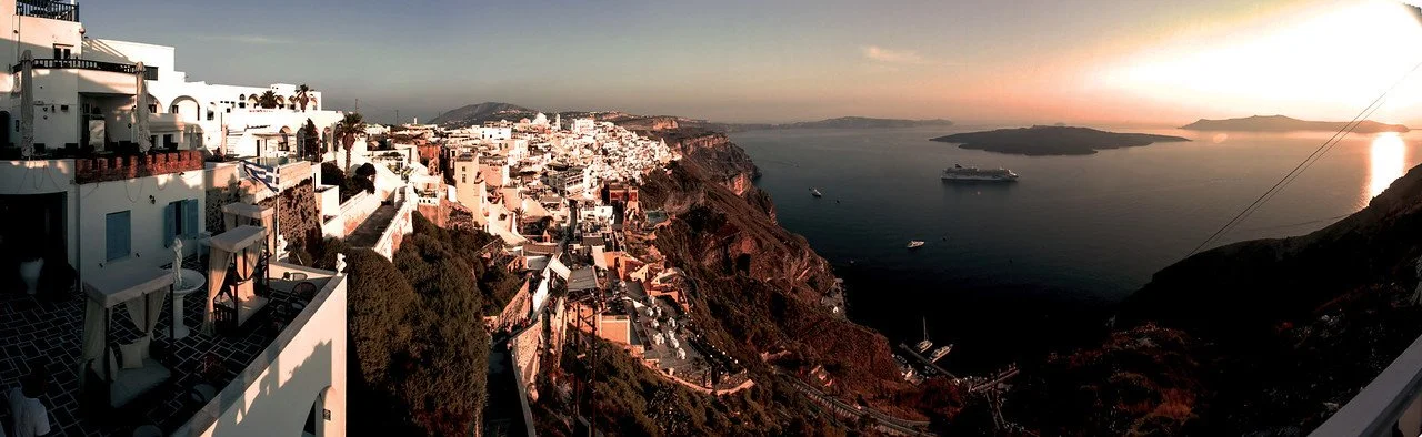 Panoramic view of a coastal town built on cliffs at sunset, with white buildings, a harbor with boats, and a cruise ship in the water.