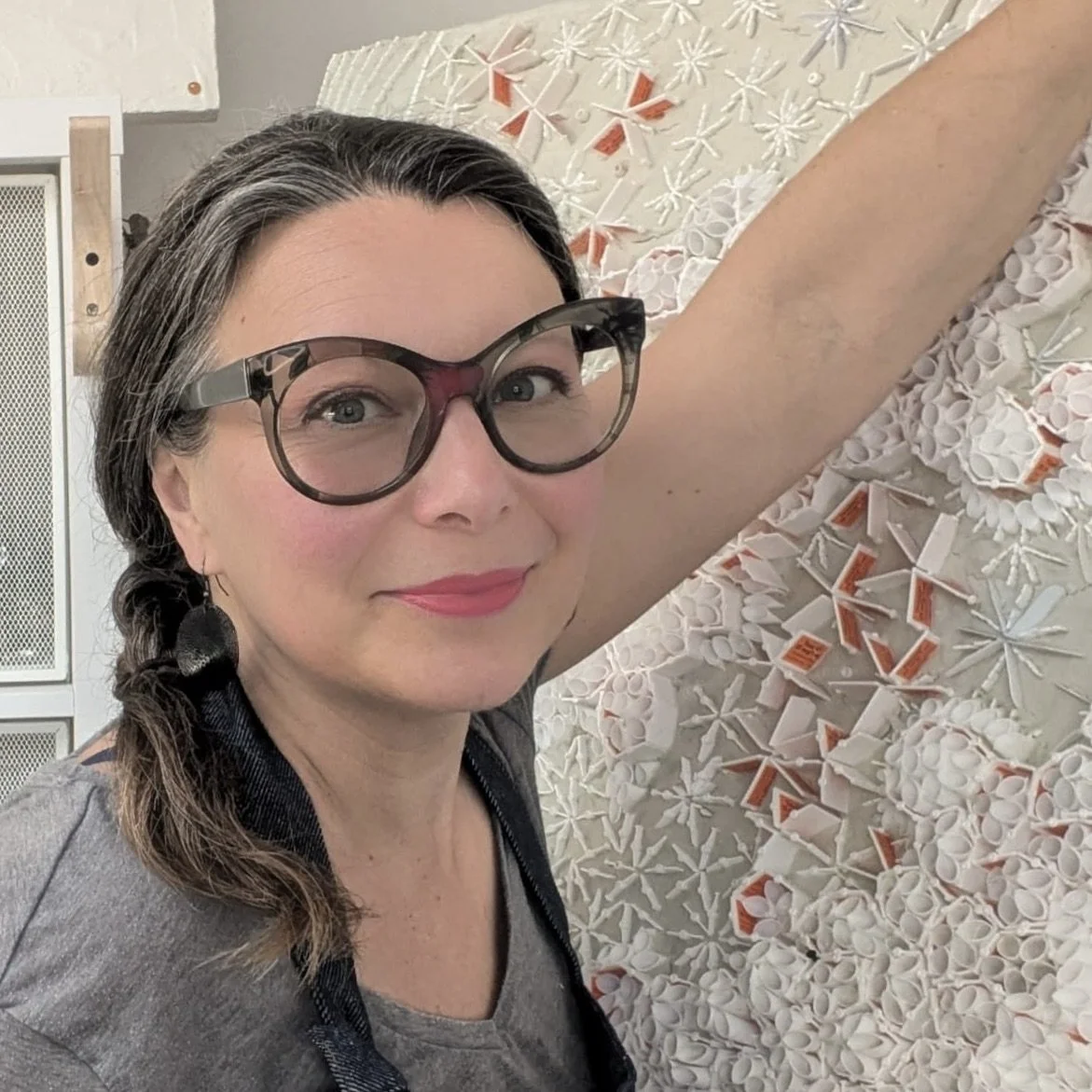 Woman with gray hair, glasses, and pink lipstick smiling and extending her arm towards a wall decorated with various white and orange geometric and floral designs.