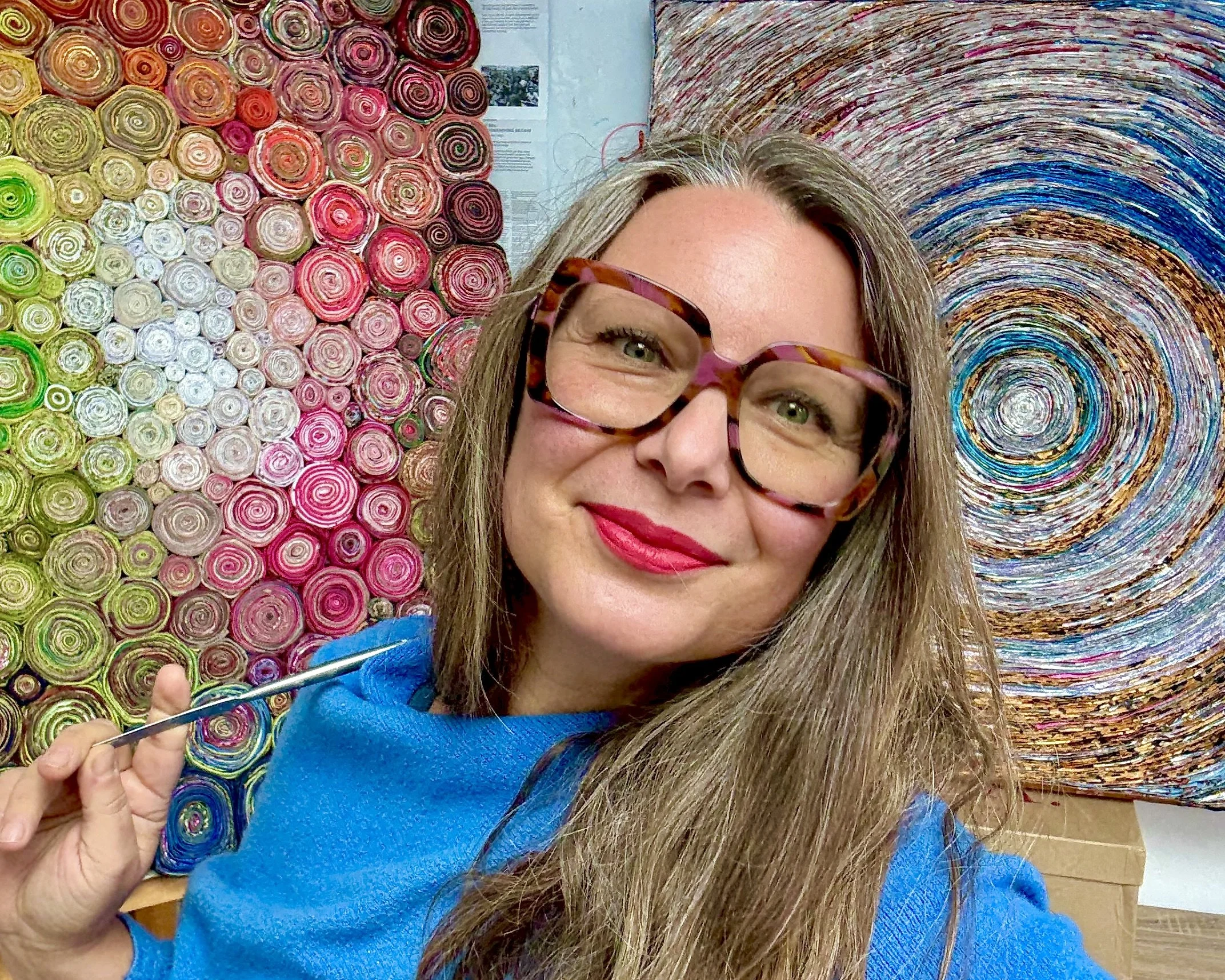 Woman with gray hair, glasses, and pink lipstick smiling and extending her arm towards a wall decorated with various white and orange geometric and floral designs.