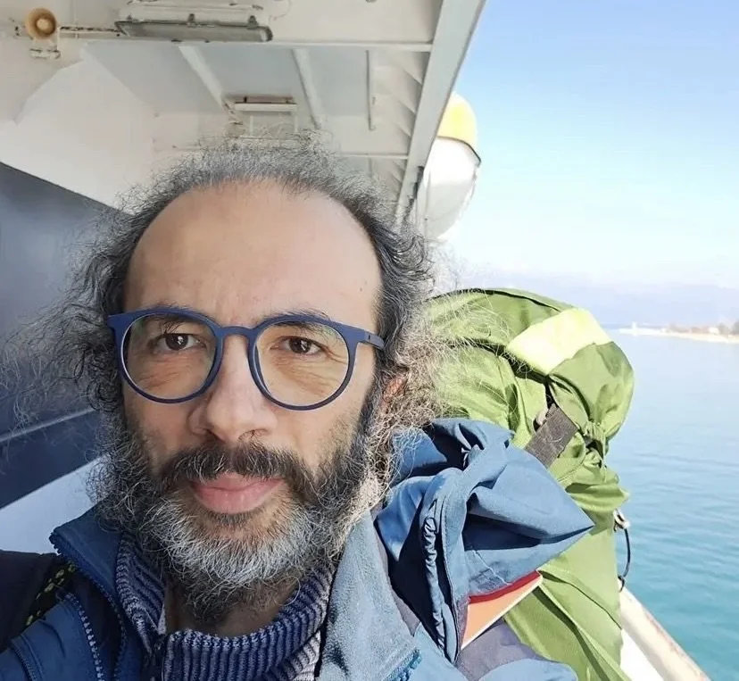 A man with glasses and a beard taking a selfie on a boat with water in the background, wearing a blue jacket and carrying a green backpack.