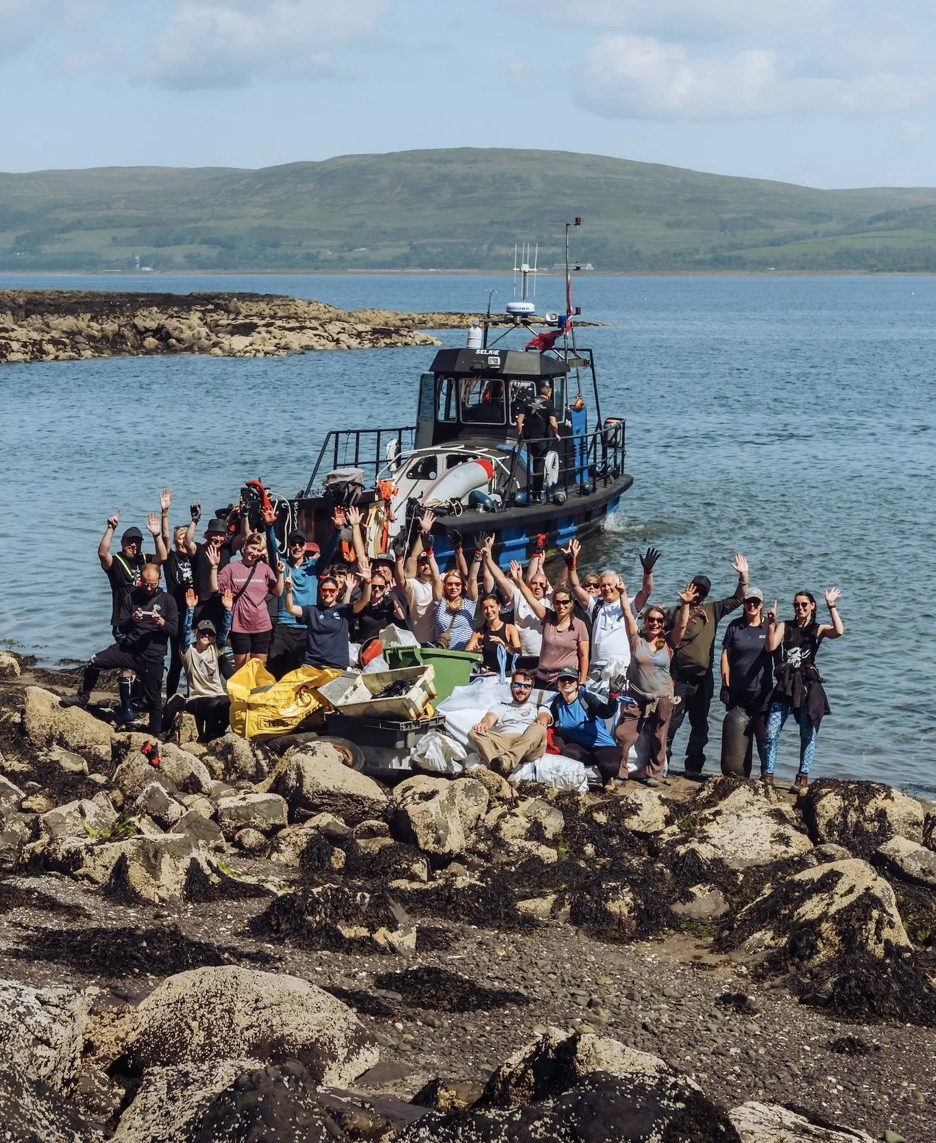 Group of people on rocky shoreline near a boat, waving and smiling, with supplies and equipment around them, during daytime with body of water and hills in background.