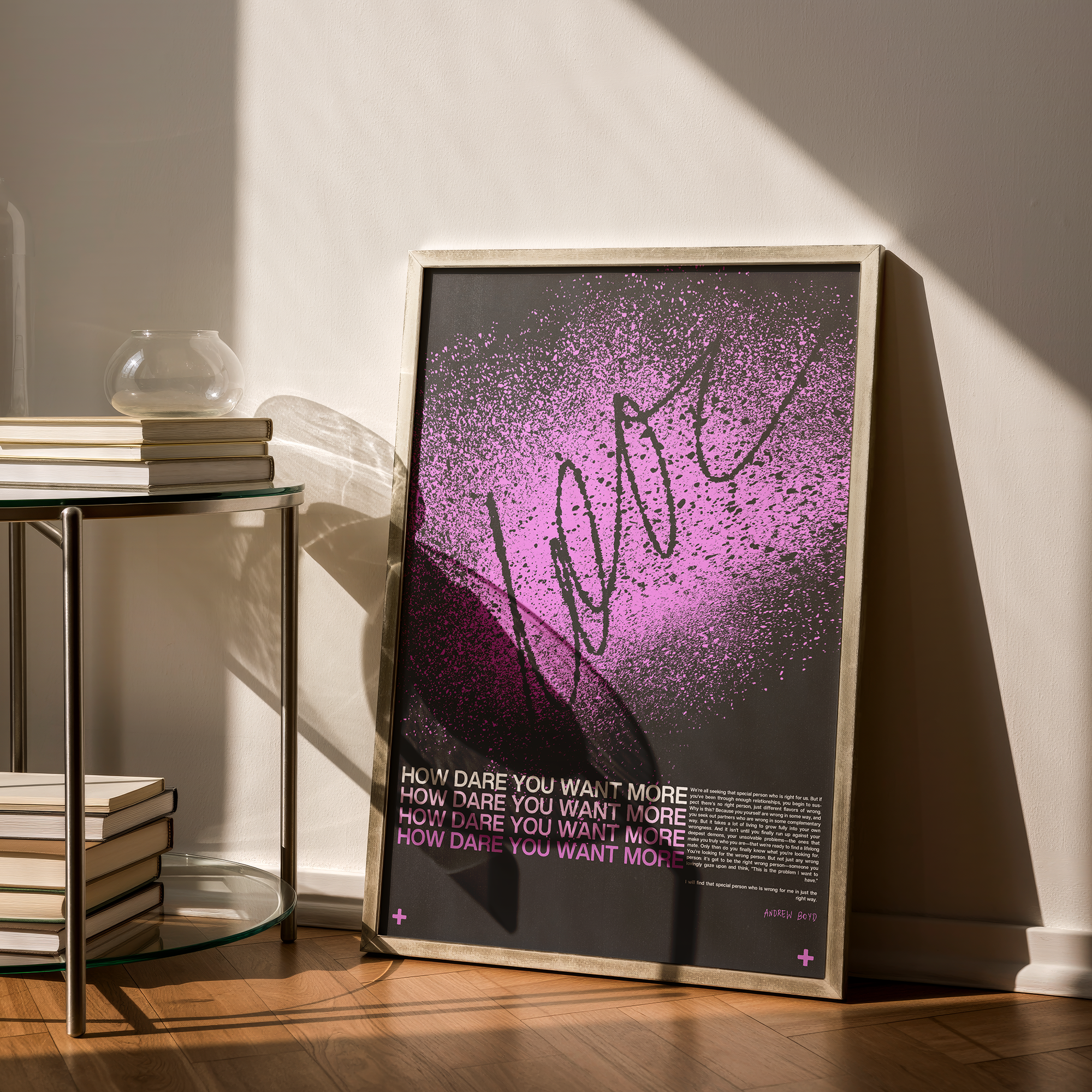 Framed artwork on a stand with pink and black text reading "HOW DARE YOU WANT MORE" repeated multiple times, placed next to a glass table with a stack of books and a glass vase, with sunlight and a shadow cast on the wall.