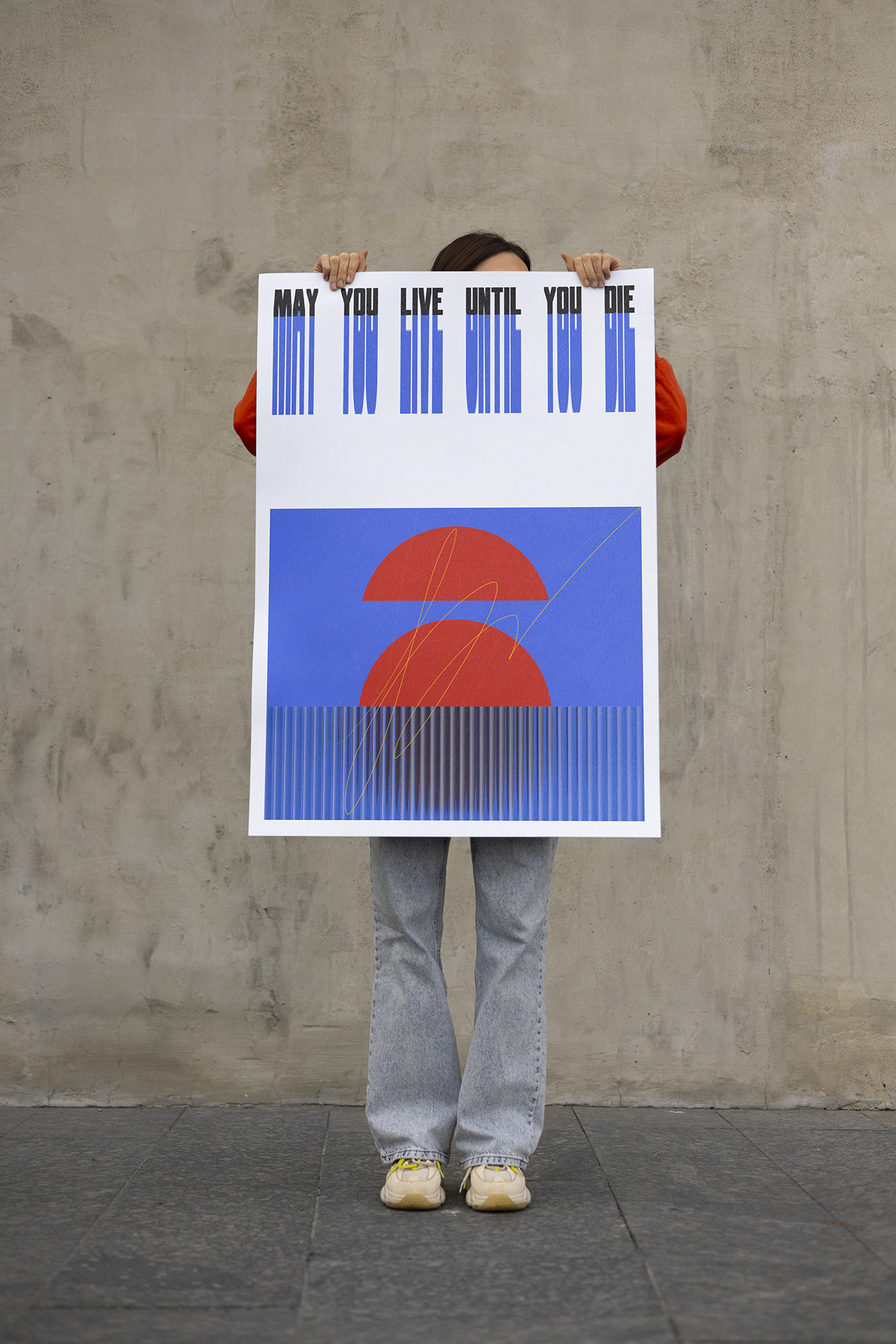 A person standing in front of a plain wall holding a large poster that reads "May you live until you die" with a colorful abstract design.