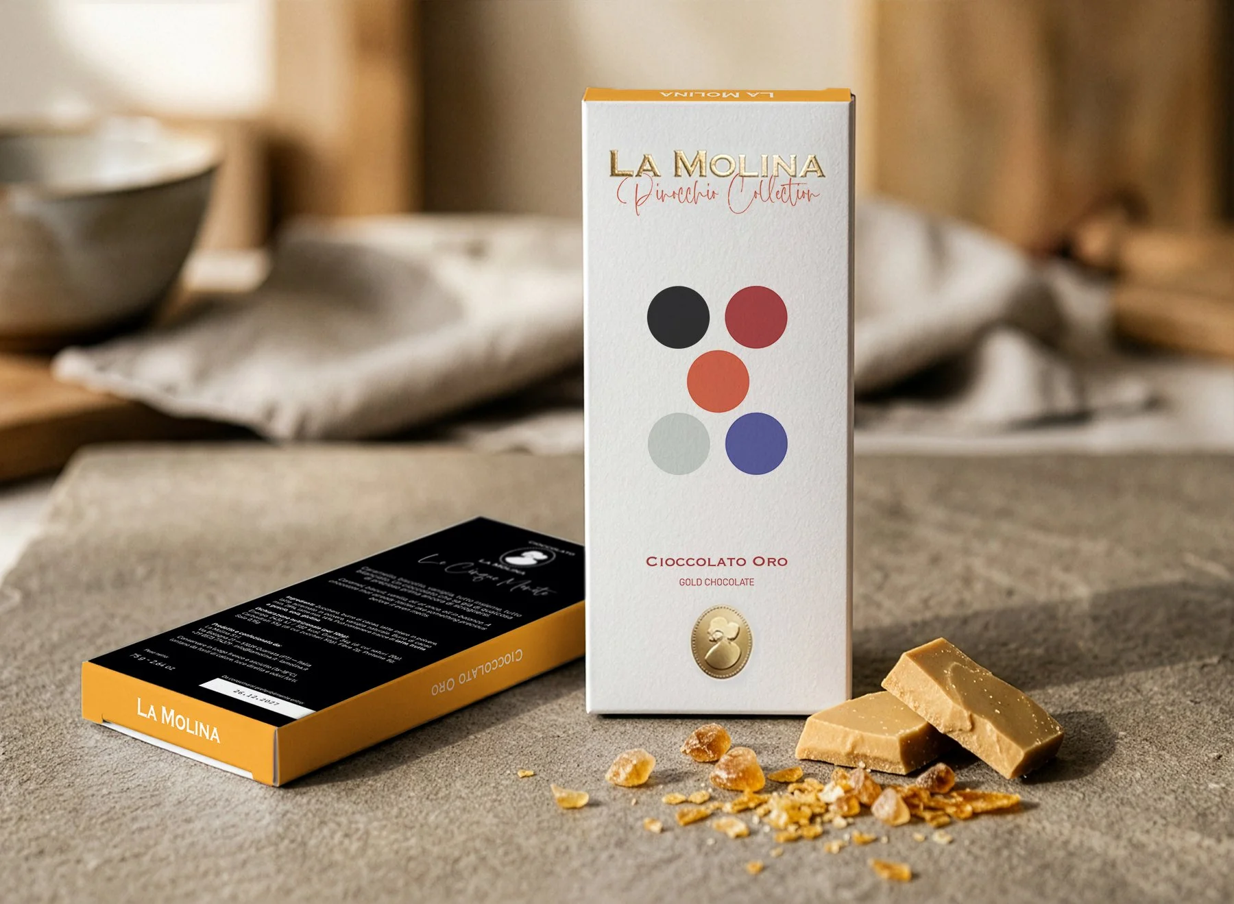 A white box of La Molina 'Pinocchio Collection' gold chocolate standing on a gray surface, with broken pieces of caramel-colored chocolate, some chunks, and scattered caramel crumbs in front. The background includes blurred kitchen items and a black rectangle with white text.
