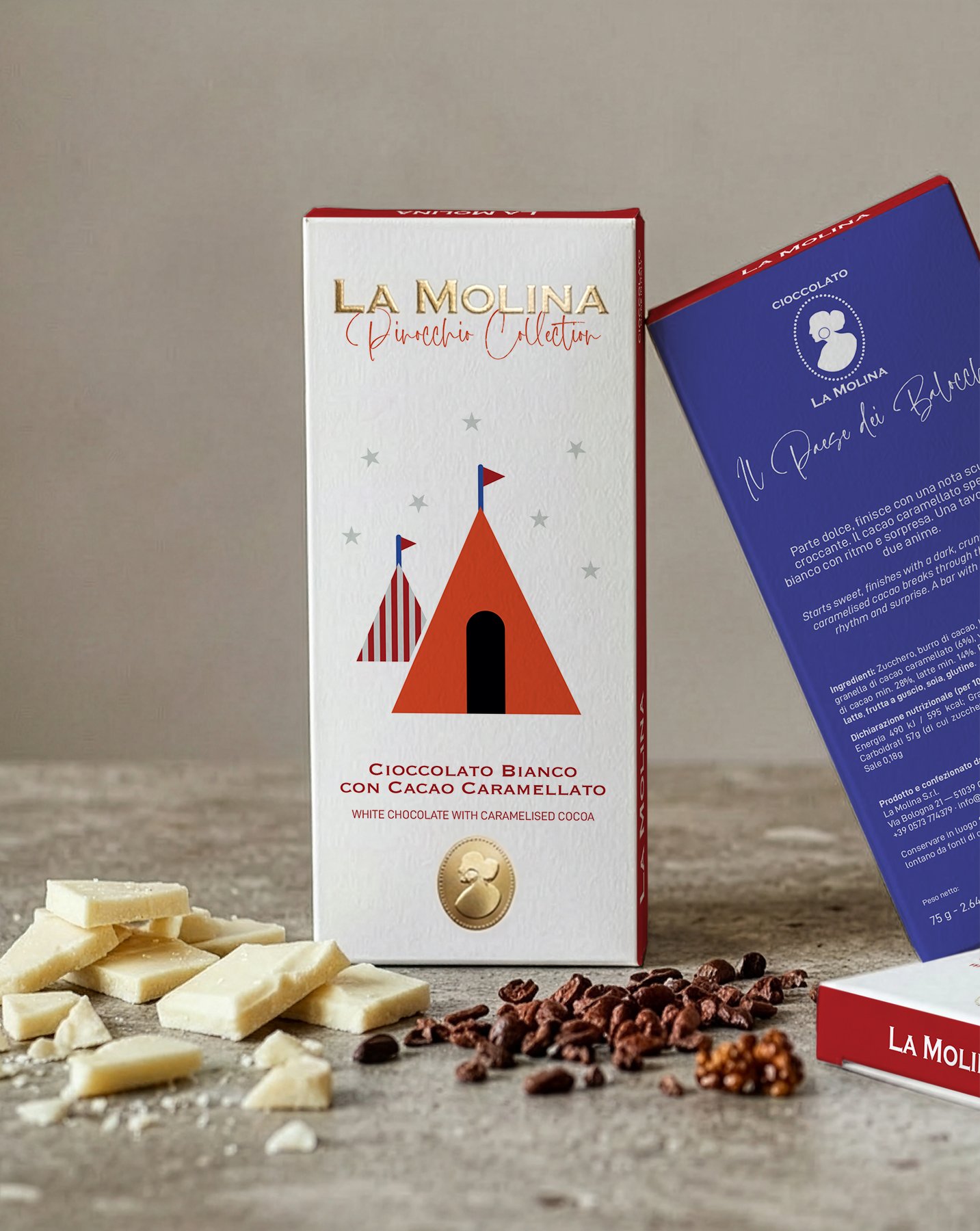 A box of La Molina Pinocchio Collection white chocolate with caramelized cocoa, with white chocolate pieces, cocoa nibs, and white chocolate shavings placed in front of it on a gray surface.