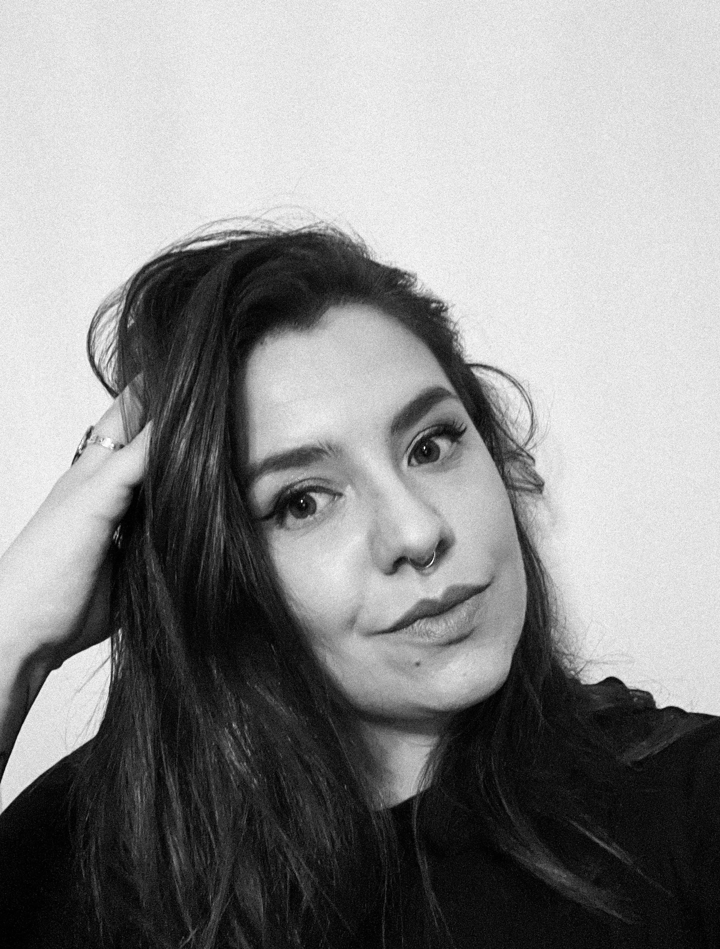 A black and white photo of Klaudia with long hair, partially visible face with prominent eyes, nose ring, posing with one hand touching their hair.