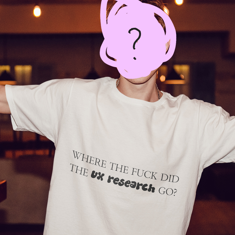 Person wearing a white T-shirt with the text "WHERE THE FUCK DID THE UX research GO?" printed on it, with arms outstretched, in a dimly lit room.