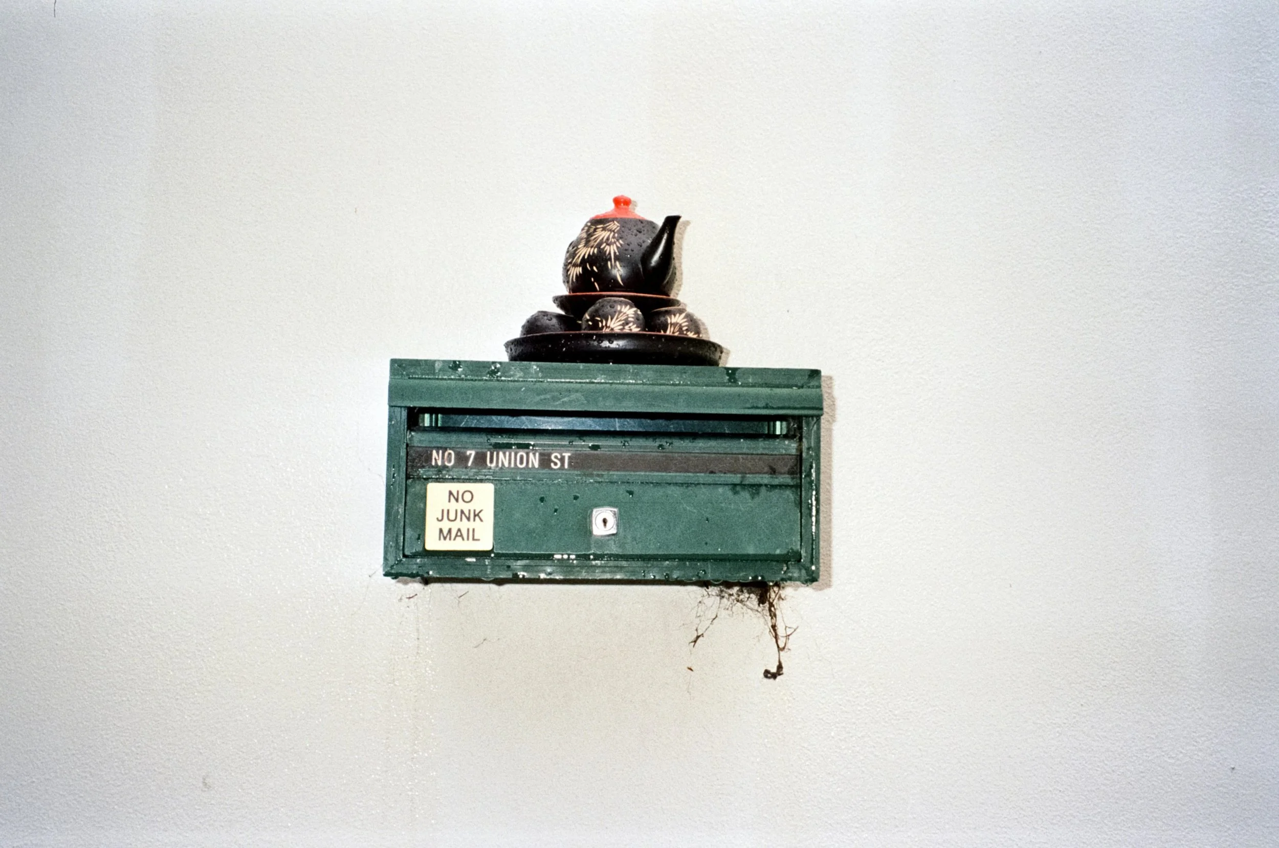 A green mailbox mounted on a wall with a stack of decorative black and white patterned teapots on top, and a small spider web hanging from the bottom.