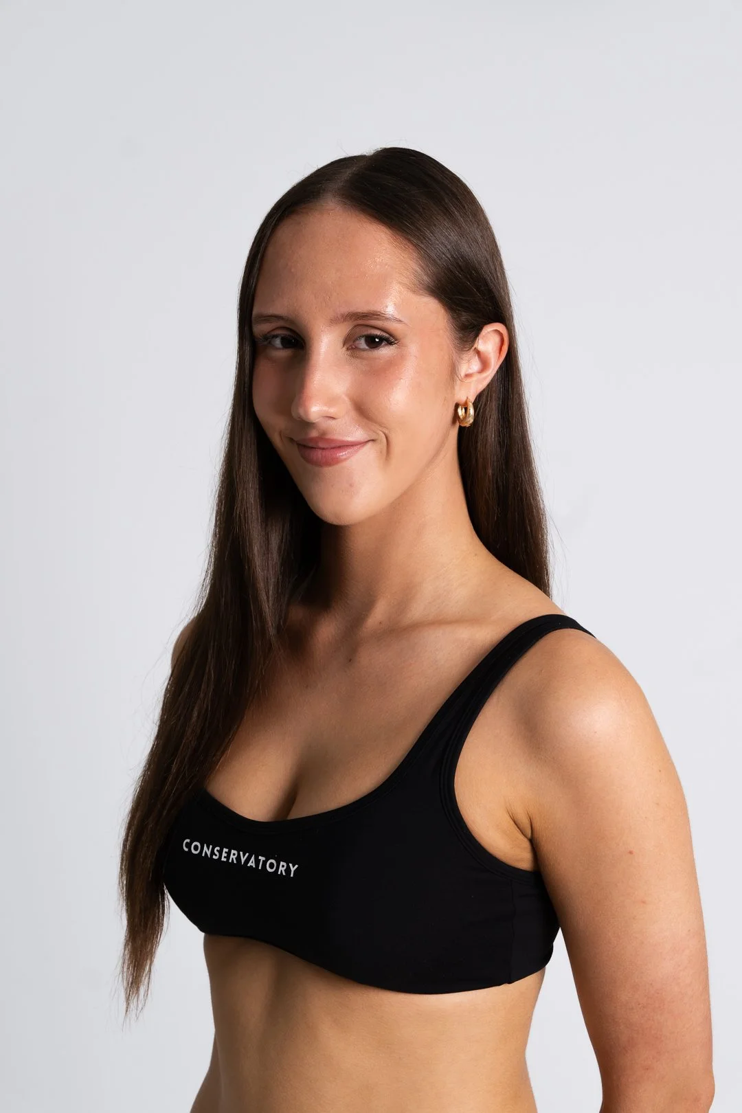 A part time dancer with long brown hair wearing a black sports bra with the word 'CONSERVATORY' written on it, smiling slightly against a plain white background.