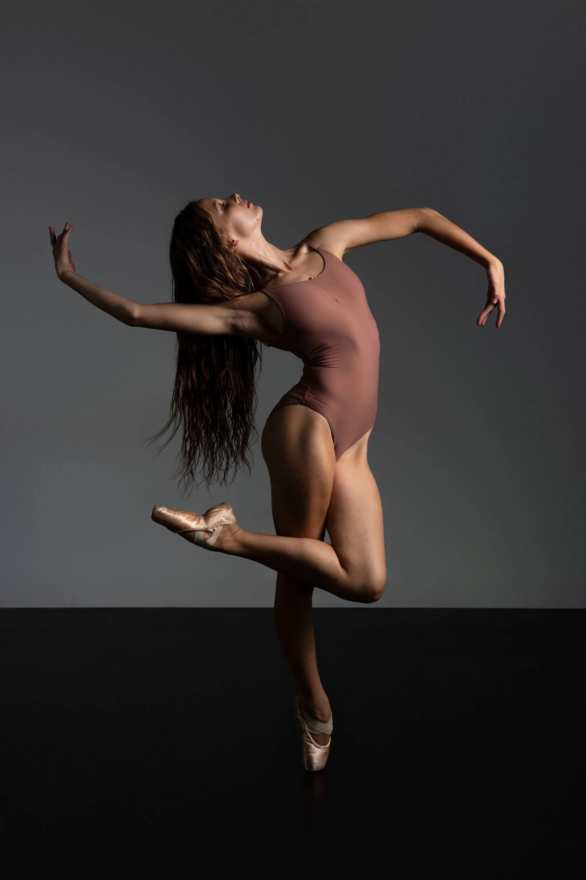 Ballet dancer in a pose with arms and one leg raised, wearing a pink leotard and pink pointe shoes, against a gray background.