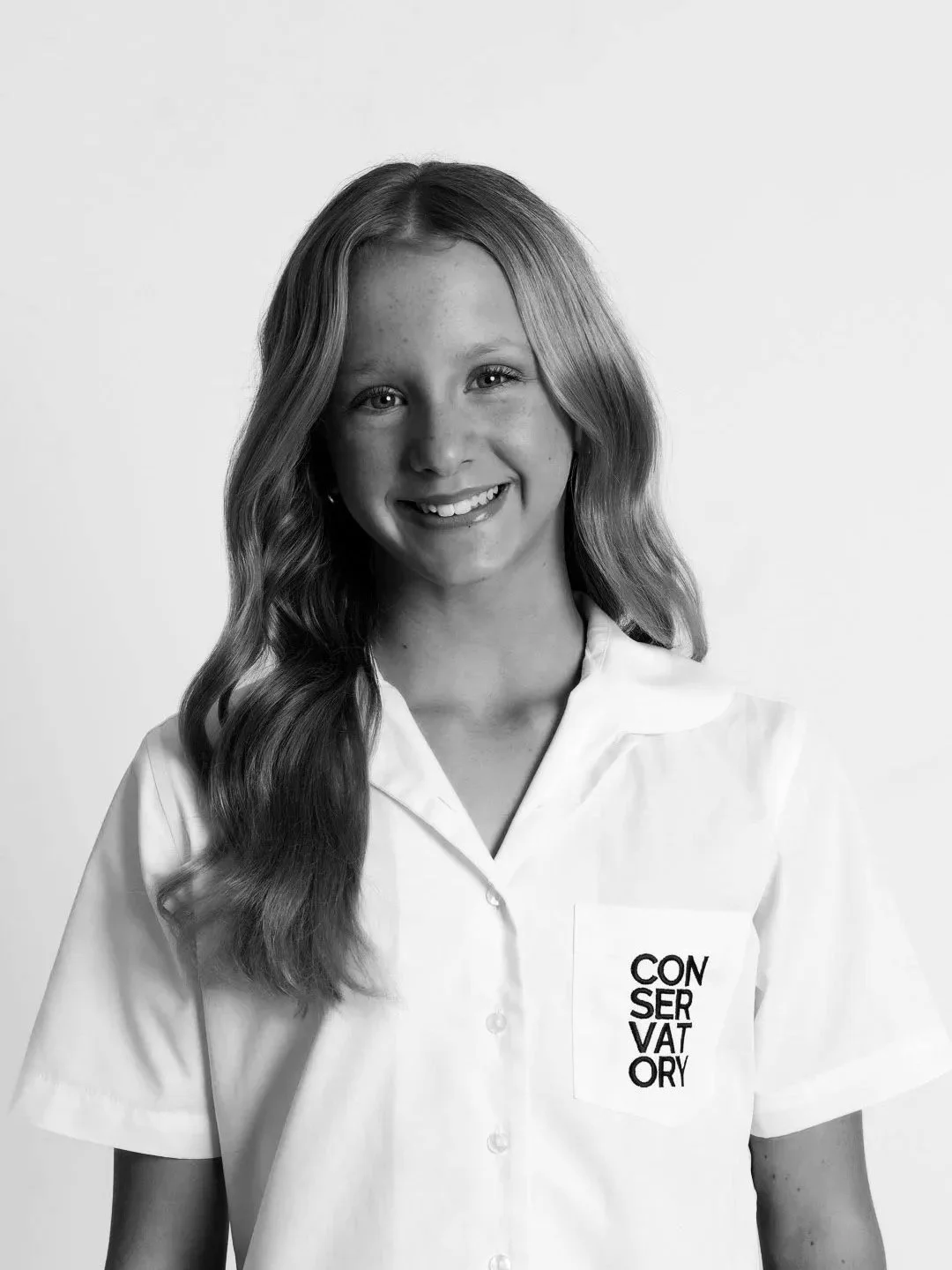 A college dancer with long wavy hair smiling in a white shirt with the word "CONSERVATORY" printed on it, against a plain background.