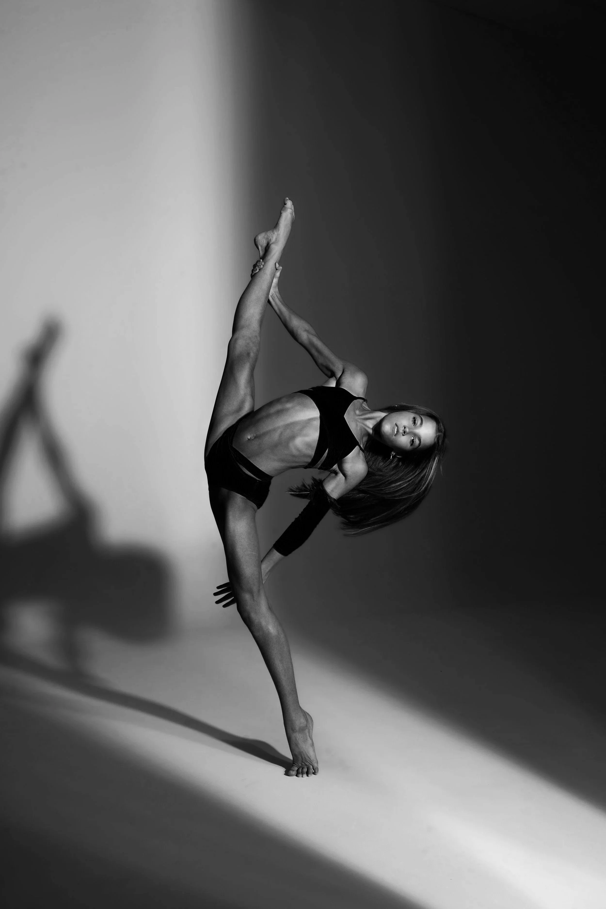 A black and white photograph of a woman in a dynamic dance pose, balancing on one foot with her other leg extended upward, and her body arched sideways with one arm extended and the other touching her head. She is wearing a sports bra and shorts, with dramatic lighting creating shadows and highlights.