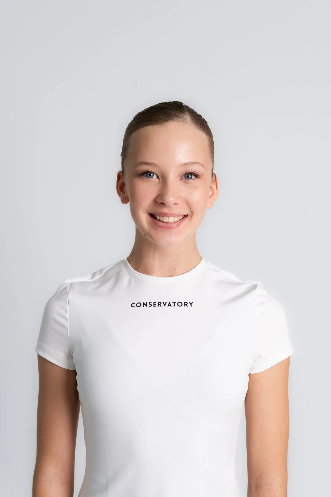A young ballet dancer with blonde hair tied back, smiling, wearing a white t-shirt with the word 'CONSERVATORY' printed on it, standing against a plain light gray background.