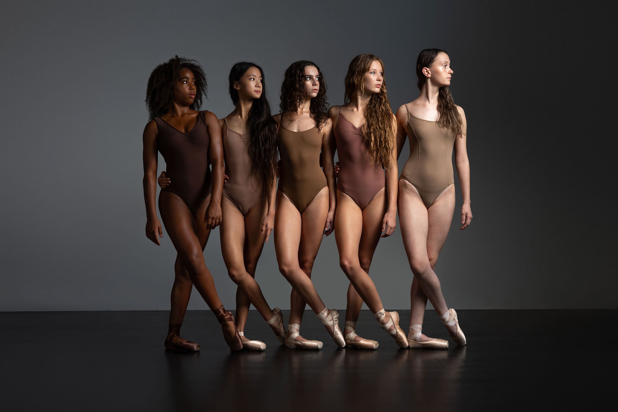 Five women in different skin tones, wearing nude-colored leotards and ballet shoes, standing in a line with arms around each other against a gray background.