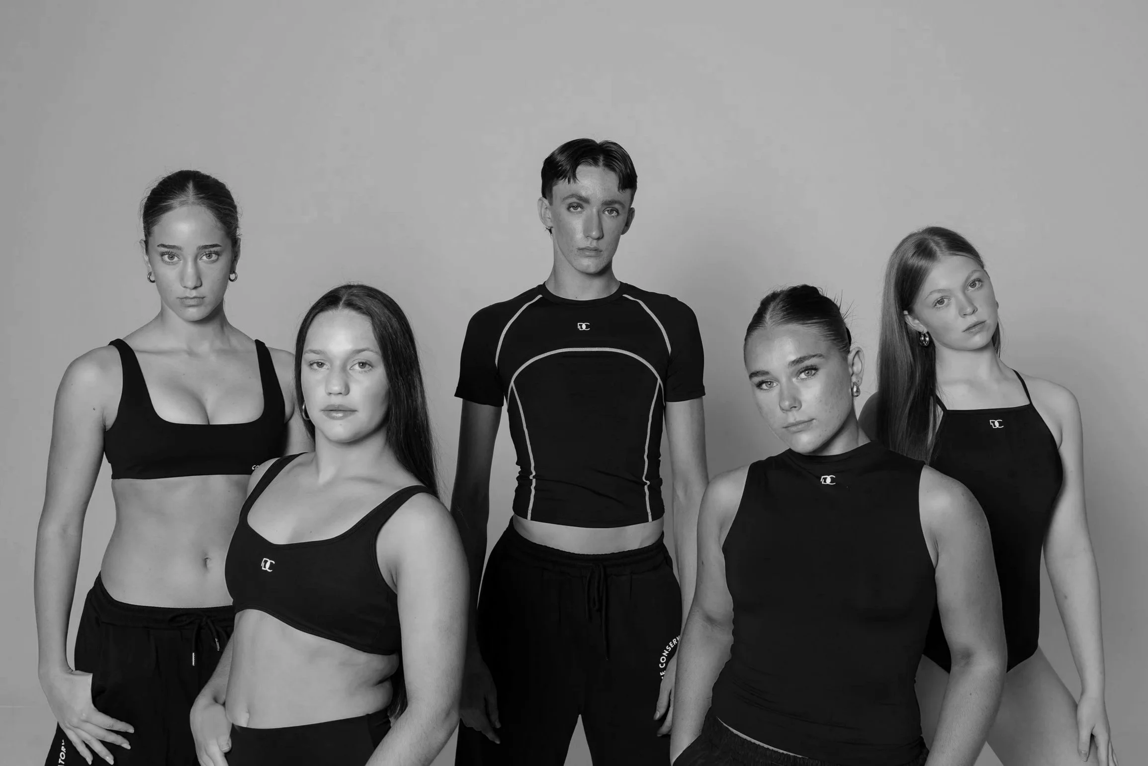 Black and white photo of four young women and one young man posing together as part time dancers, wearing athletic clothing, against a plain background.