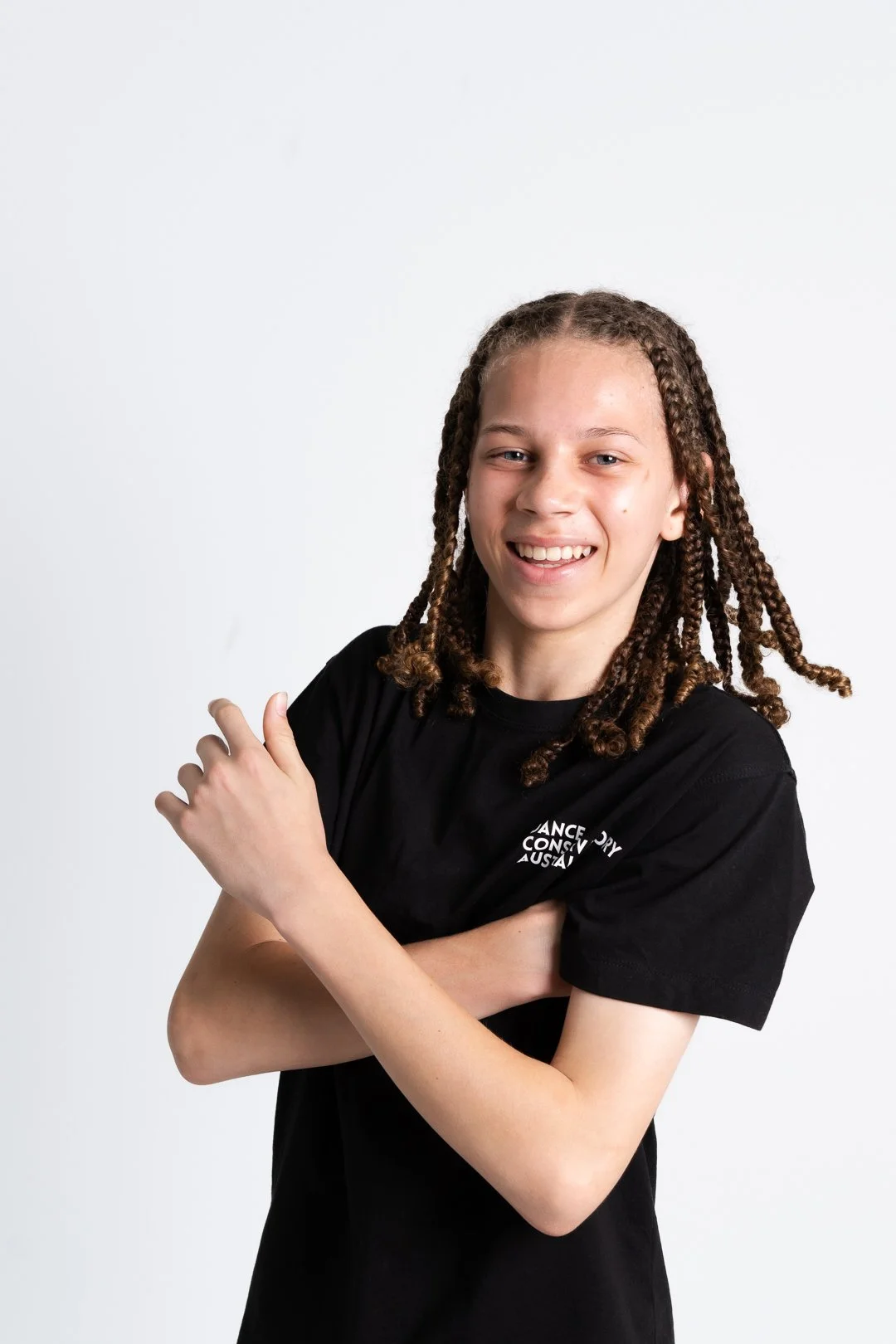 A young college dancer with long brown curly hair, smiling and crossing their arms, wearing a black T-shirt.