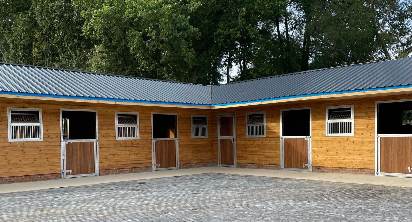 A wooden stable building with multiple horse stalls, each having a door with a small window, a brick base, and a metal roof.