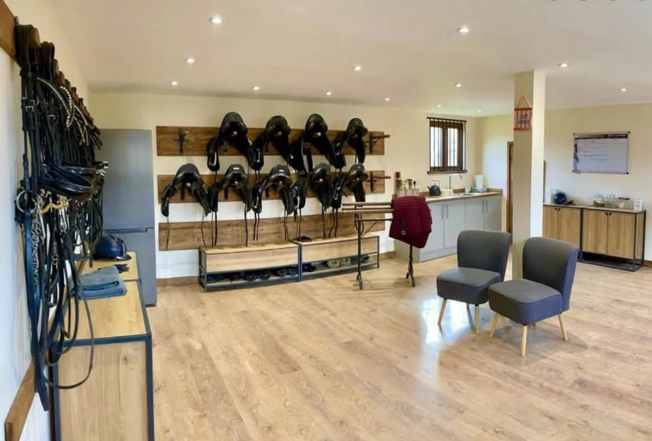 A cozy tack room with saddles and bridles organised on hooks, a small sitting area with two chairs, and a kitchenette with countertops and cabinets in the background.