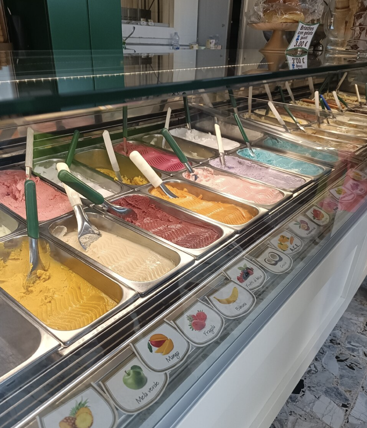 Vetrina di gelati artigianali di vari colori e gusti, con cucchiai e spatole, in una gelateria.