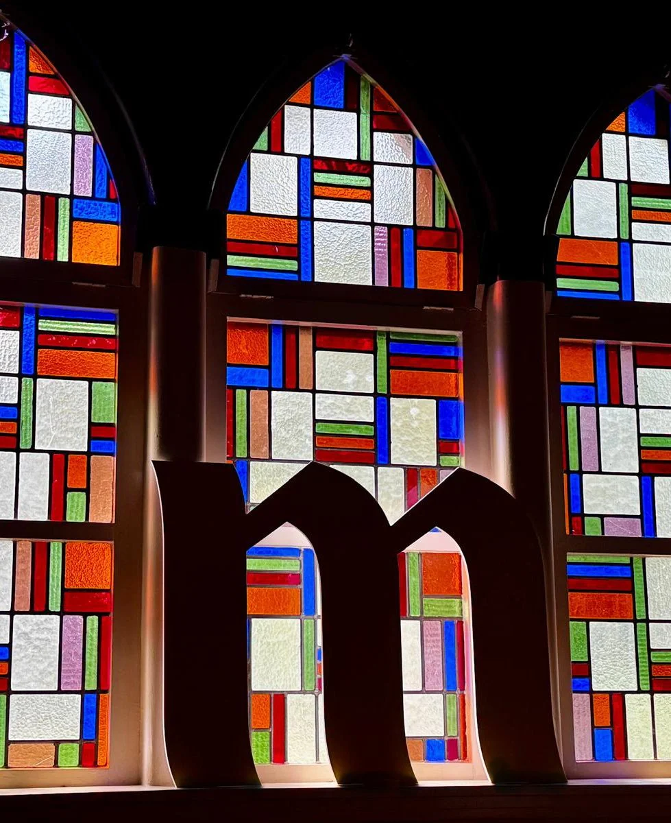 Kleurrieve glas-in-loodramen met geometrische patronen in een kerk, met een zwarte decoratieve letter M op de voorgrond.