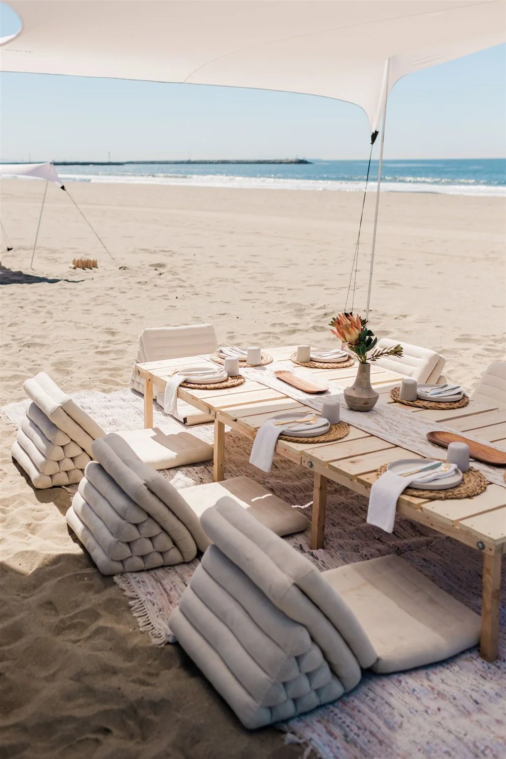 Beach picnic with white cushions tablescape white linens pink protea under shade canopy