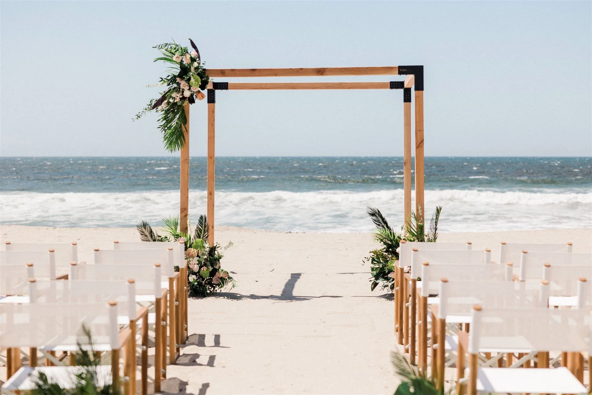 Wedding on the beach in California with square arch chuppah aisle white chairs and tropical florals