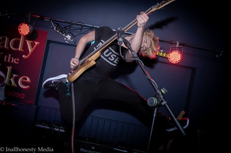 Musician with blonde hair playing an electric guitar on stage under red stage lights.