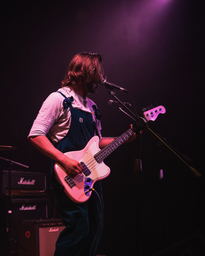A person performing on stage with a pink bass guitar and singing into a microphone, with Marshall amplifiers in the background, under purple stage lighting.