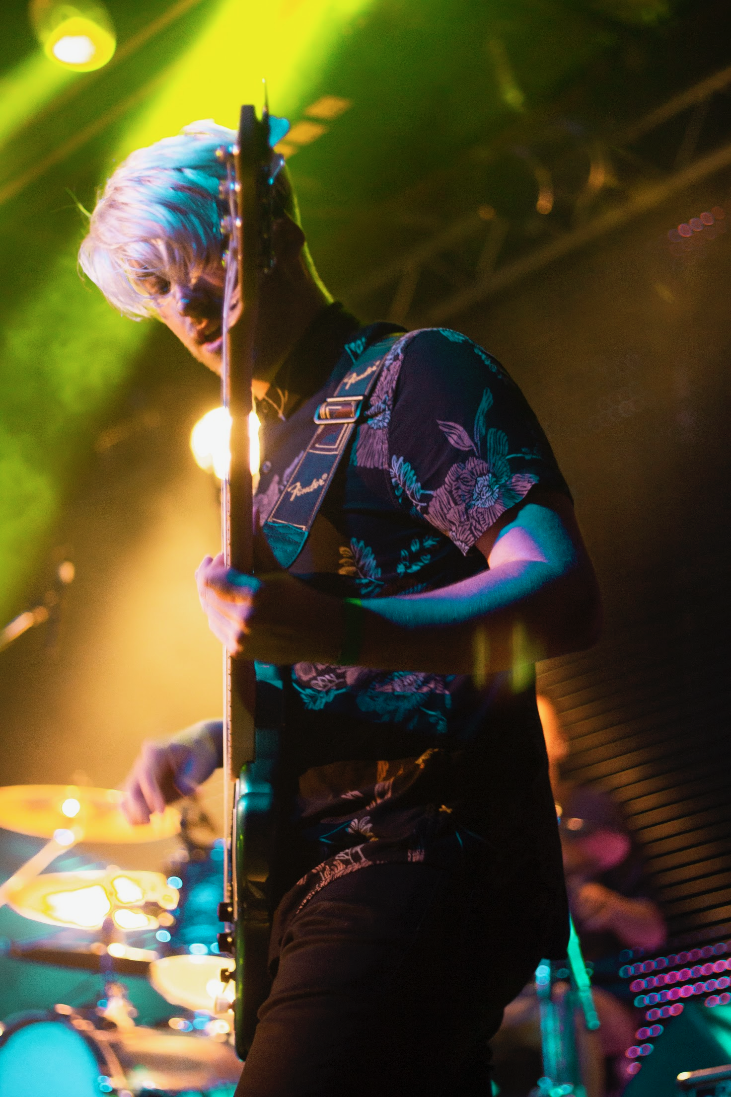 A person with blond hair playing an electric guitar on stage with colorful lighting and a drummer visible in the background.