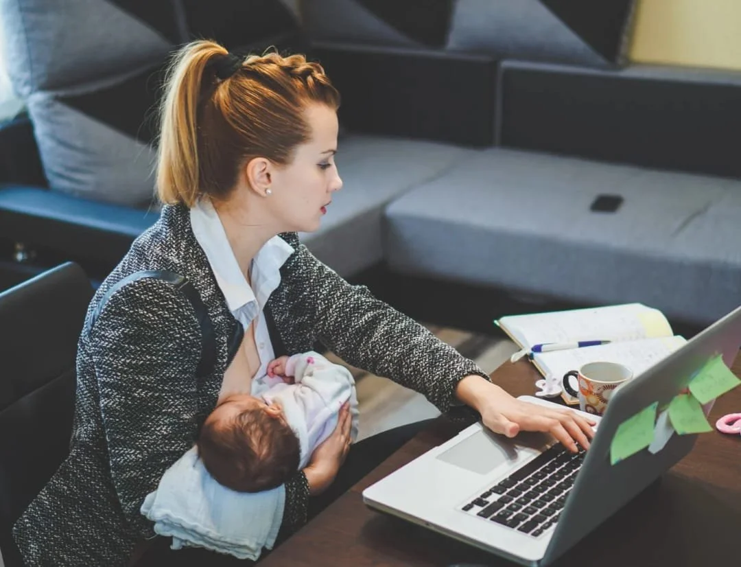 Una donna con i capelli rossi legati in una coda lavora al computer mentre allatta un neonato con una maglietta bianca, in un ambiente domestico.