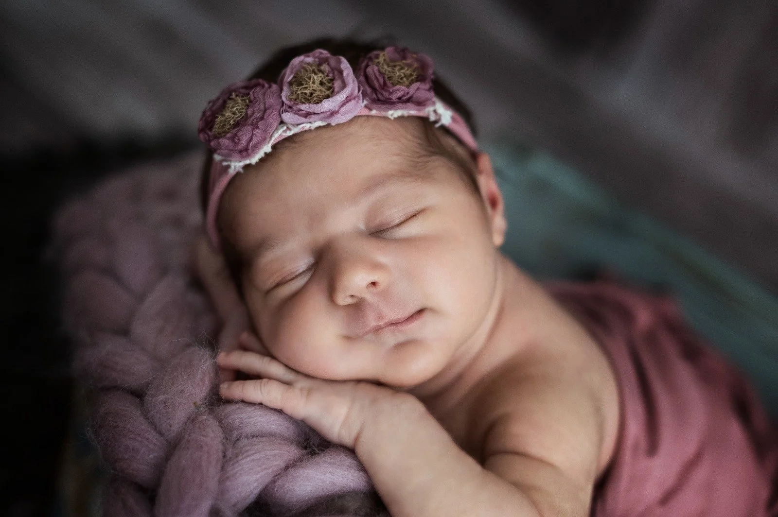 Neonato che dorme con una fascia di fiori rosa in testa, appoggiato su un morbido cuscino viola.