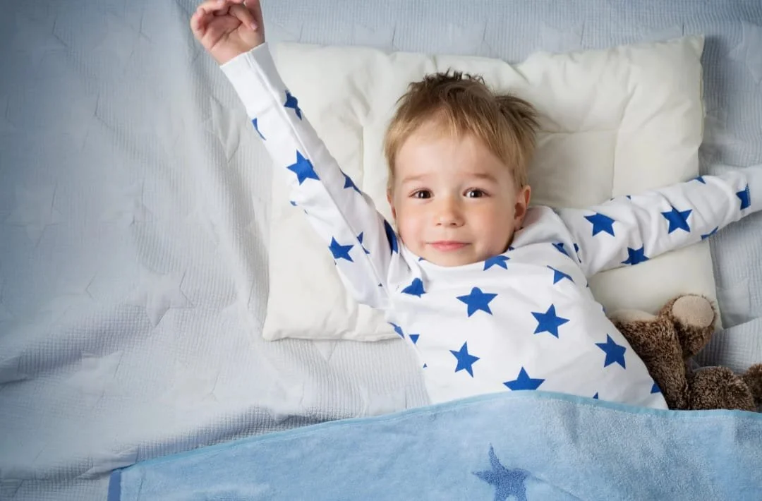 Un bambino sdraiato nel letto con una maglietta a stelline blu, che sorride e saluta con una mano, con un orsacchiotto di peluche accanto.