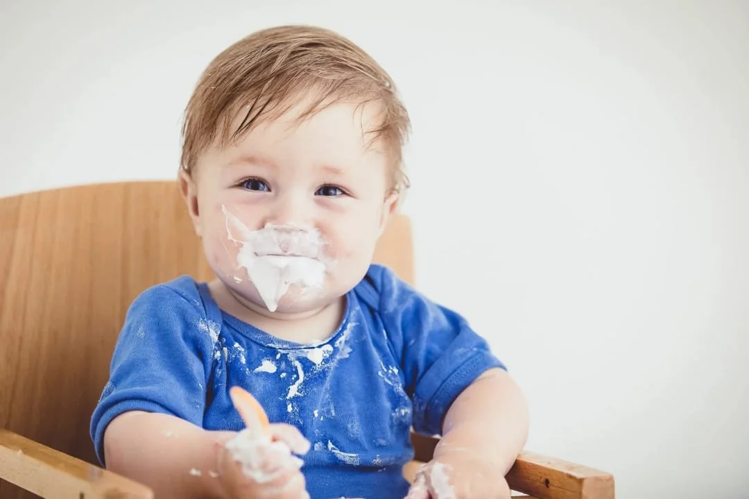 Bambino seduto in una sedia di legno, con la faccia e le mani sporche di panna, indossa una maglietta blu, sorride e tiene in mano un dito sporco di panna.
